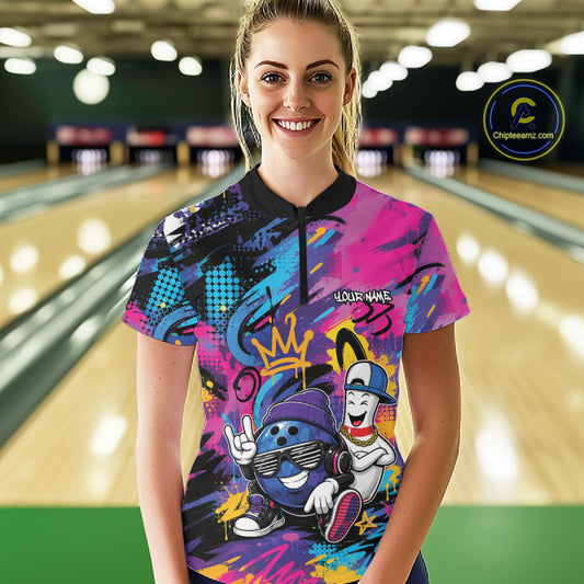 A woman stands in a bowling alley wearing a short-sleeve quarter zip bowling shirt with a vibrant multicolor graffiti-style graphic featuring cartoon bowling characters. The background shows blurred bowling lanes and lights.