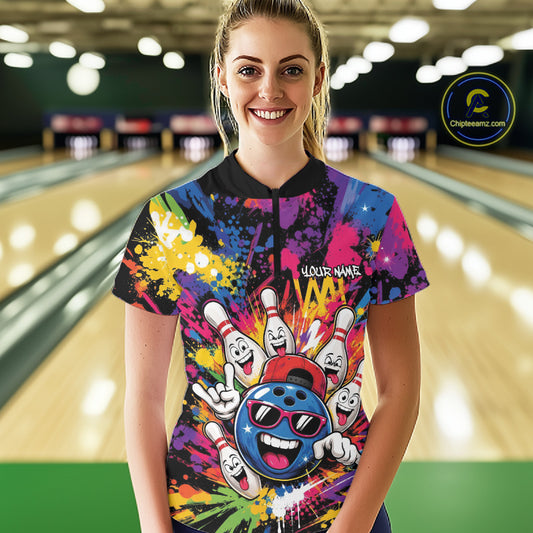 A woman stands in a bowling alley wearing a colorful short-sleeve bowling-themed graphic quarter zip shirt features a vibrant multicolor paint splatter design with cartoon bowling pins, bowling balls, and a blue bowling ball wearing sunglasses.
