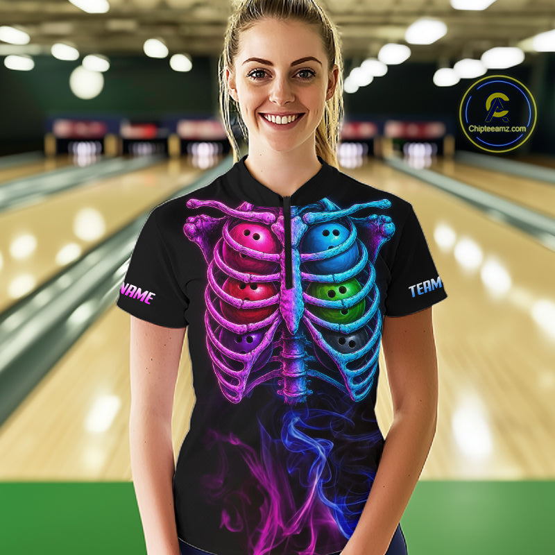 A woman stands in a bowling alley wearing a black short-sleeve shirt with a colorful graphic of a rib cage made of bowling balls and purple smoke details. The background shows blurred bowling lanes.