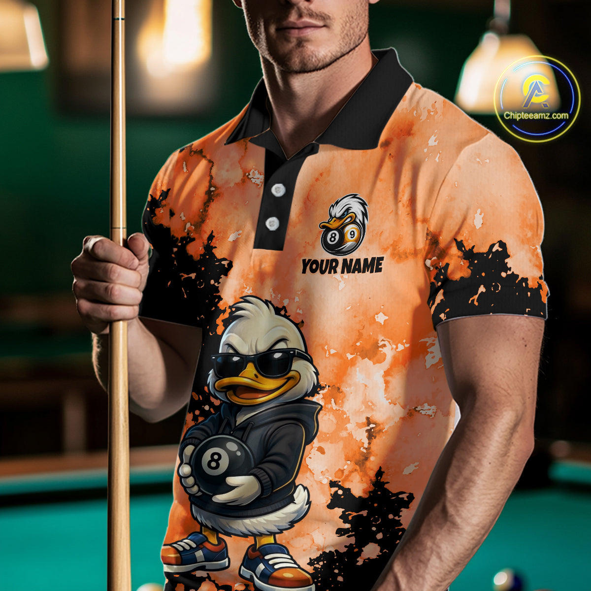 A man wearing a custom orange and black short-sleeve polo shirt with a cartoon duck graphic holding an 8-ball, holding a pool cue in a dimly lit pool hall. 