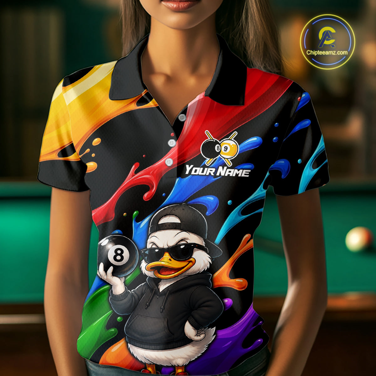 A woman wearing a short-sleeve polo shirt with a colorful abstract paint-splatter design and a cartoon duck character holding an 8-ball. The shirt has a black collar and the text 'YOUR NAME' on the chest