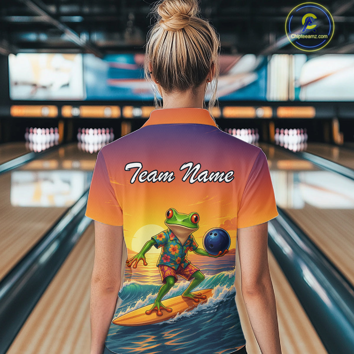A person standing in a bowling alley wearing a short-sleeve collared shirt from the back, featuring a colorful gradient from orange to purple with a large graphic of a frog surfing on a wave while holding a bowling ball. The back of the shirt has the text 'Team Name' printed on it.