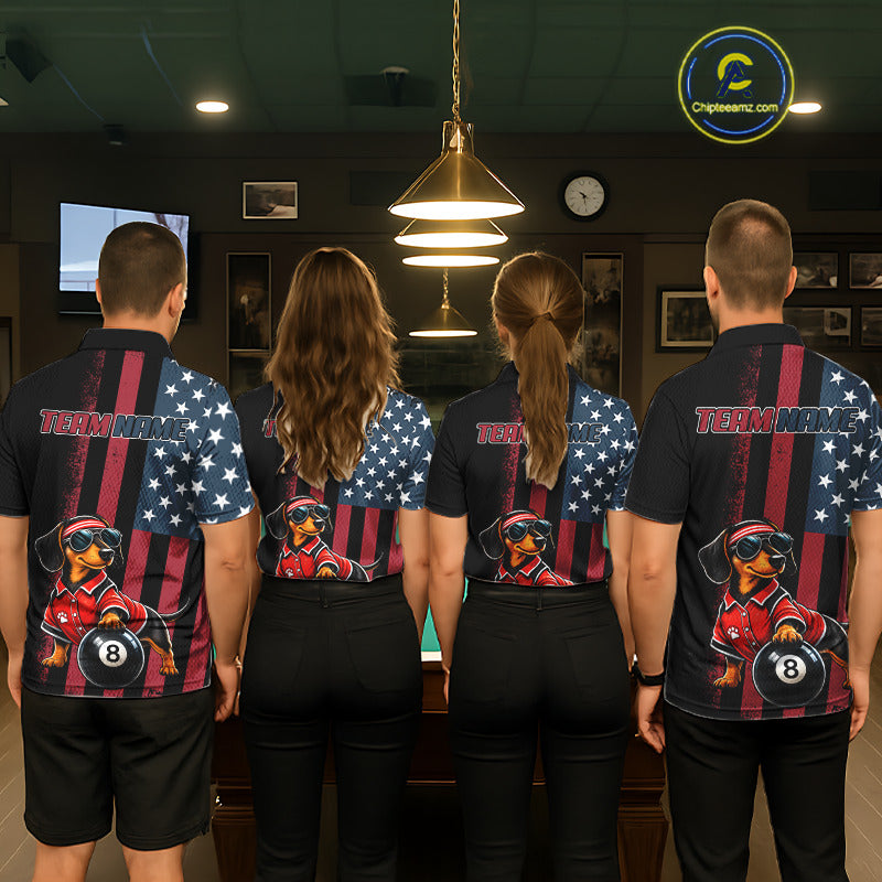 Four people standing with their backs to the camera in a dimly lit bar or lounge, wearing matching short-sleeve collared shirts with a patriotic American flag design, red and white vertical stripes, white stars on a blue field, and a large cartoon dachshund billiards graphic on the back.