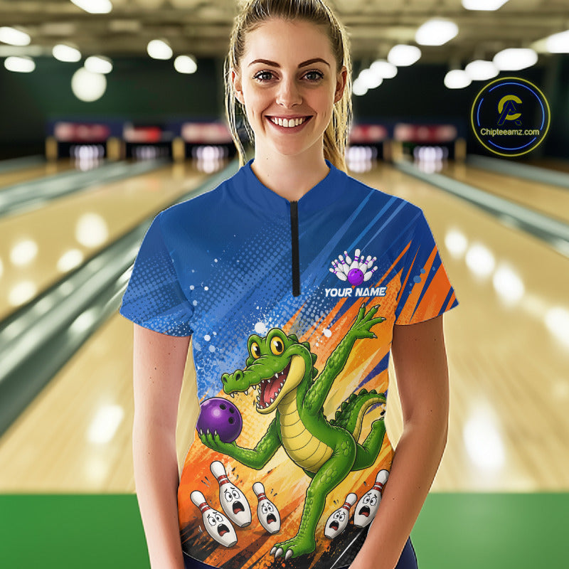 A woman stands in a bowling alley wearing a short-sleeve bowling jersey with a colorful cartoon alligator holding a purple bowling ball, orange and blue graphic panels, and a black zipper at the neck. 