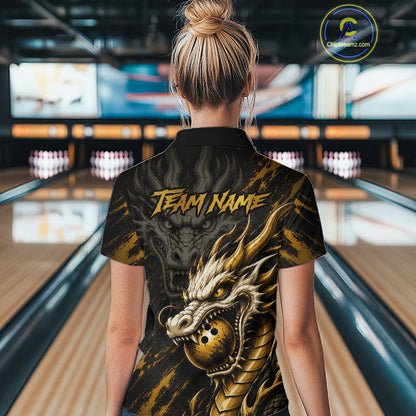 A woman viewed from behind wearing a black short-sleeve polo shirt with a large gold dragon graphic and the text 'TEAM NAME' on the back, standing in a bowling alley.