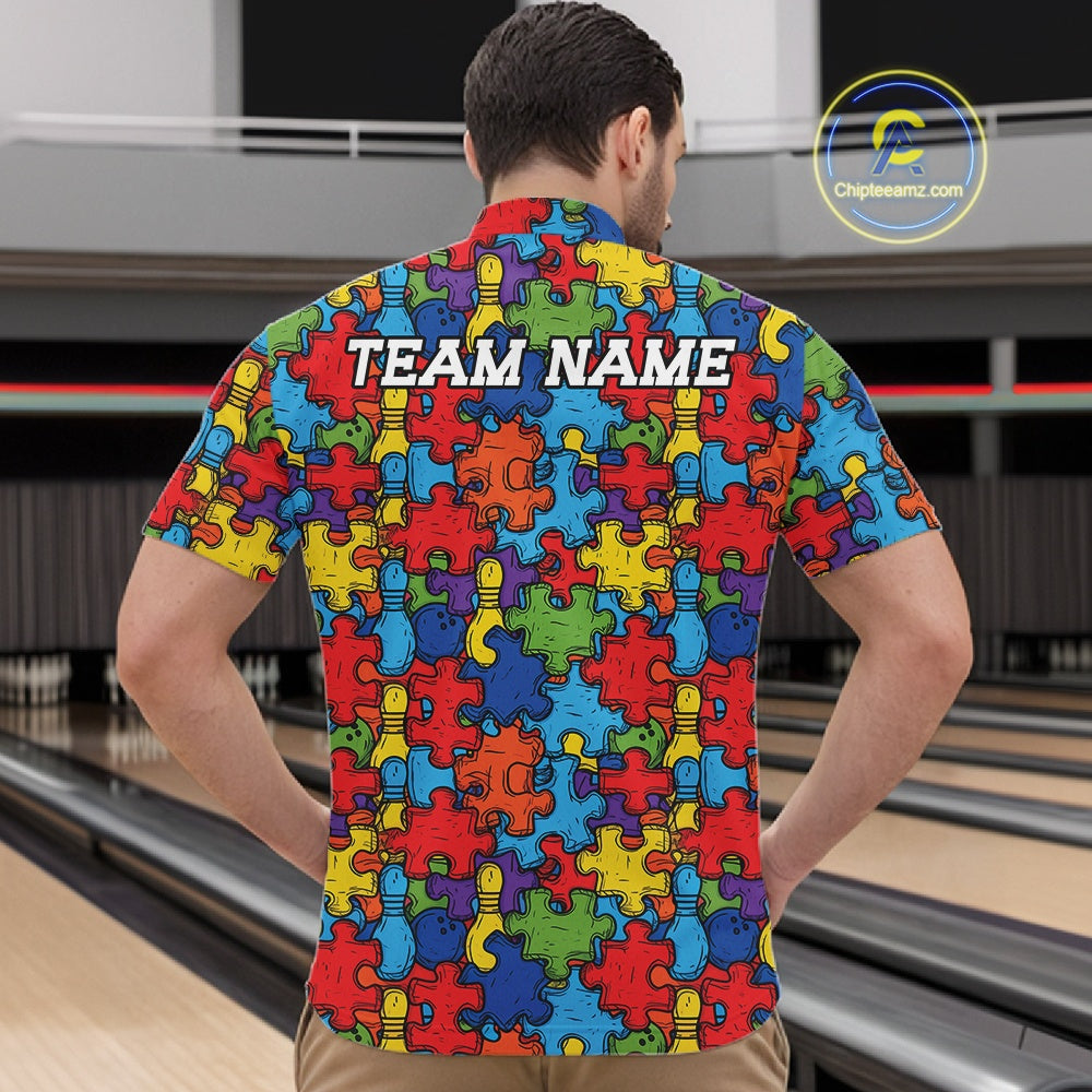 A man viewed from behind wearing a short-sleeve  shirt with a colorful puzzle-piece pattern and the text 'TEAM NAME' printed across the upper back. Bowling alley lanes are visible in the background