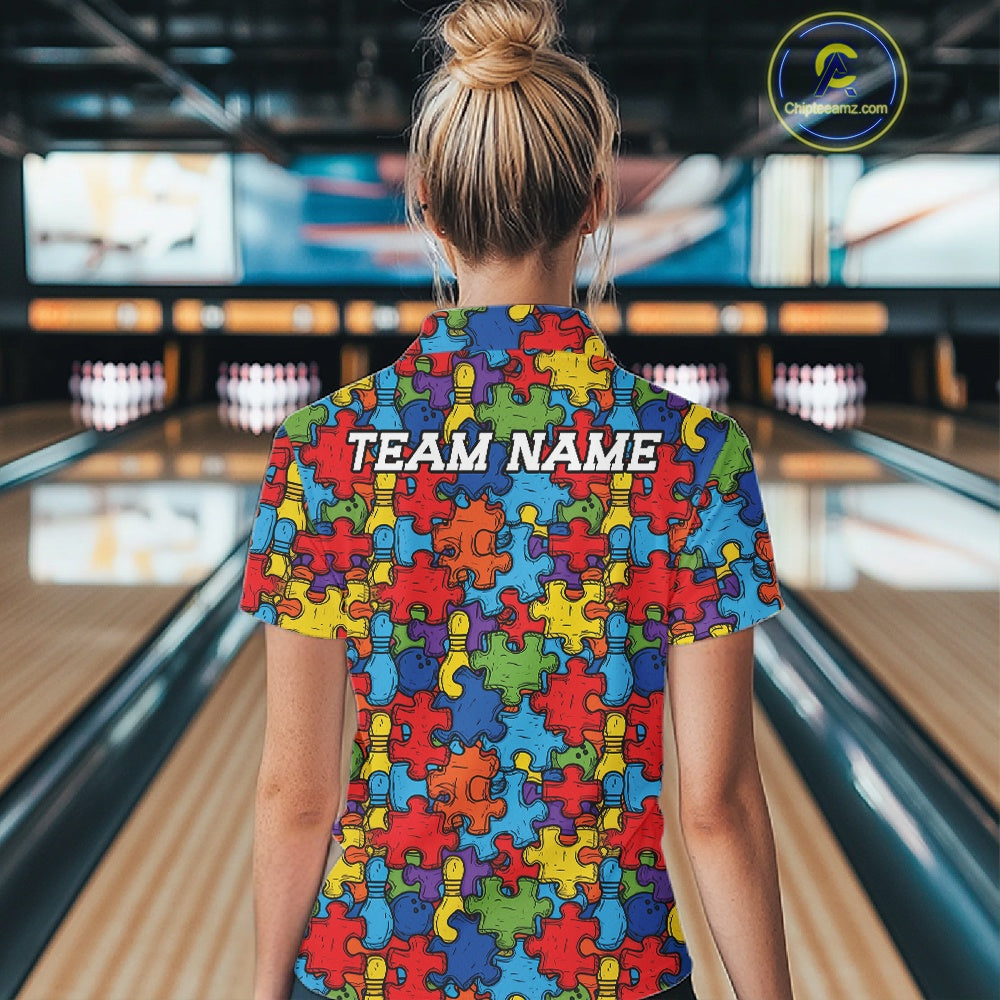 A woman viewed from behind wearing a short-sleeve collared shirt covered in a colorful puzzle-piece pattern, standing in a bowling alley. The shirt has the words 'TEAM NAME' printed across the back.
