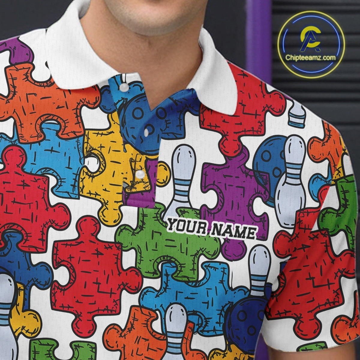 A close-up of a person wearing a white polo shirt covered in a colorful puzzle-piece pattern, with bowling pins integrated into some pieces. The shirt has a white collar and a customizable name printed on the chest.