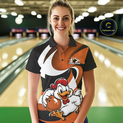 A woman stands in a bowling alley wearing a custom printed short-sleeve polo shirt. The shirt features a black and orange design with a large cartoon rooster graphic holding a bowling ball on the front.