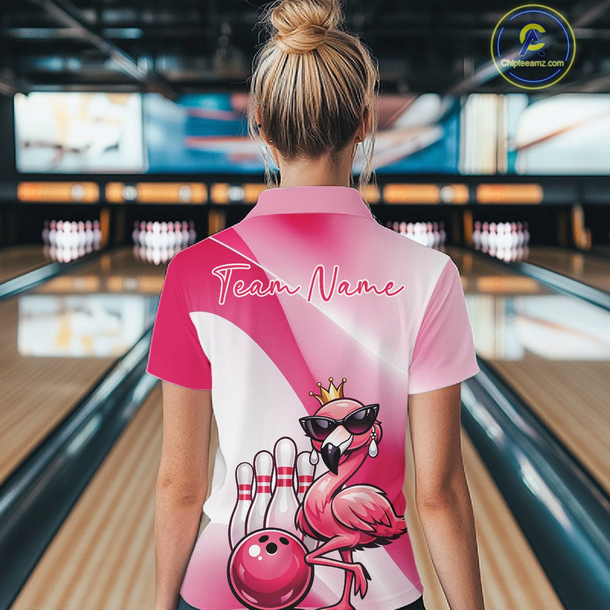 Back view of a female model in a bowling venue wearing a pink quarter-zip shirt featuring a crowned flamingo and bowling ball artwork.