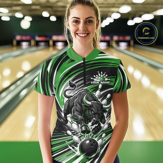 A woman stands in a bowling alley wearing a short-sleeve quarter zip bowling shirt with a green, black, and white graphic design featuring a charging bull and bowling graphic. The background shows blurred bowling lanes and lights.