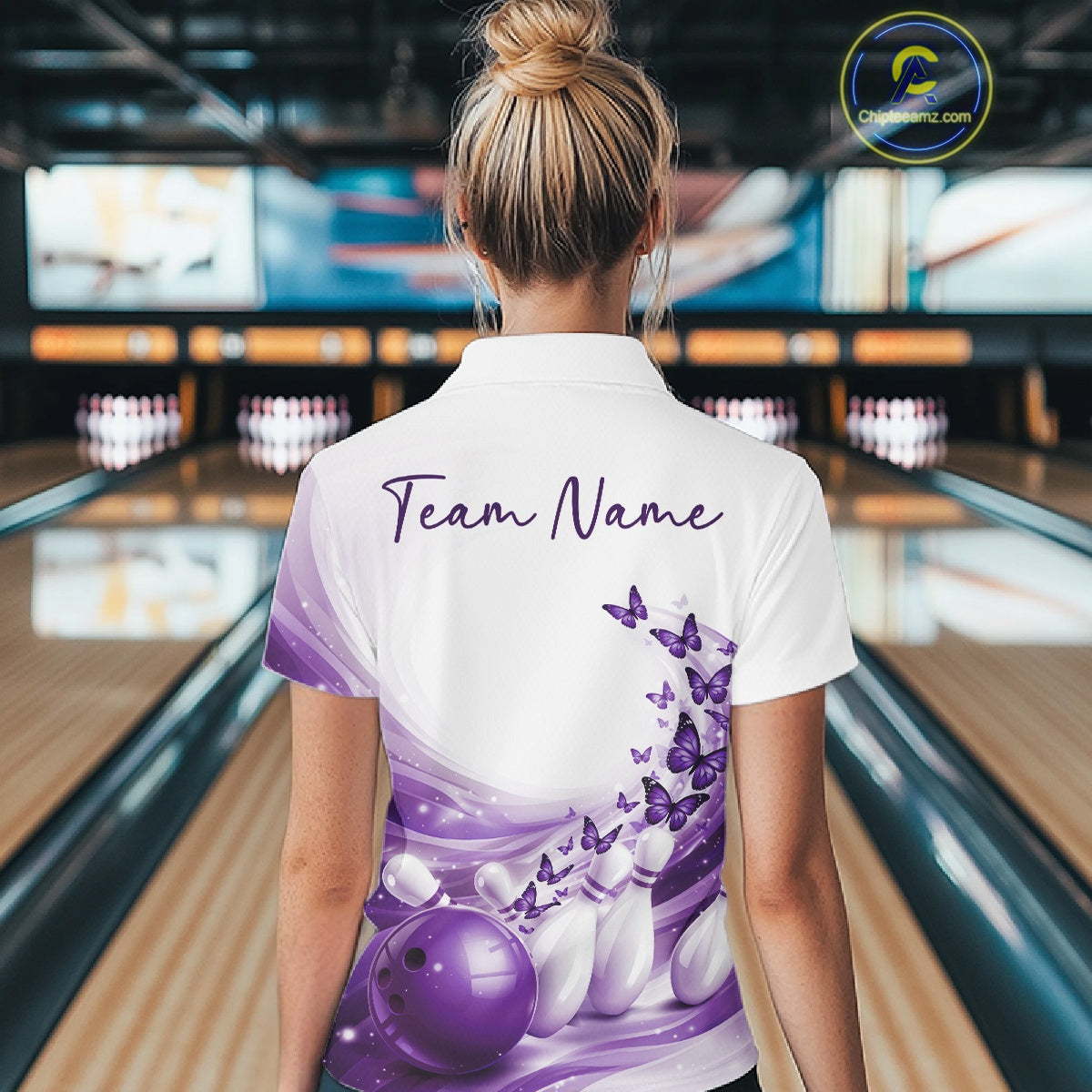 Female model wearing a white and purple bowling polo with butterfly artwork and bowling graphics, back view at the bowling lane