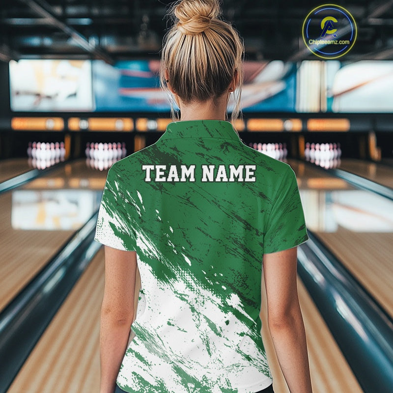 Back view of a female model in a bowling alley wearing a bowling quarter-zip shirt featuring a white paint splatter distressed sportswear design on a green shirt.
