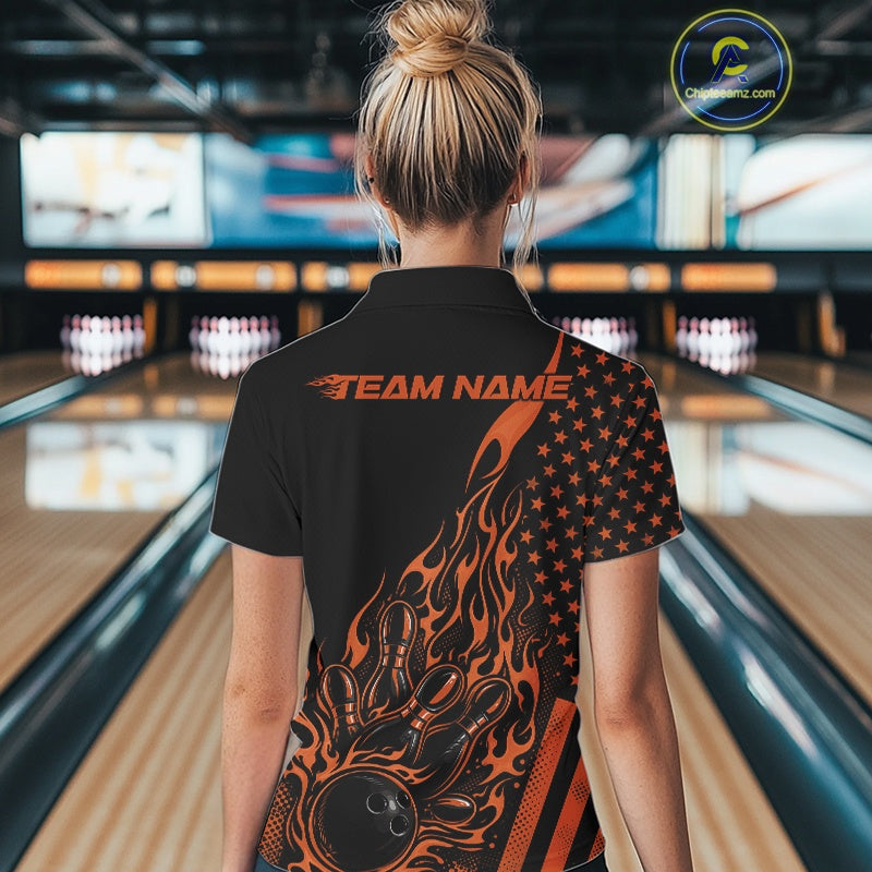 Back view of a female model in a bowling center wearing a black polo shirt featuring an orange flaming bowling ball and patriotic pin artwork.