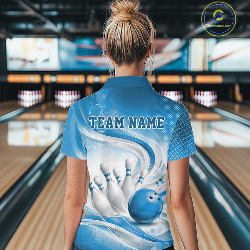 Female model standing in a bowling lane showing the back of a white and blue bowling quarter-zip shirt featuring abstract wave lines and a glossy bowling ball striking pins.