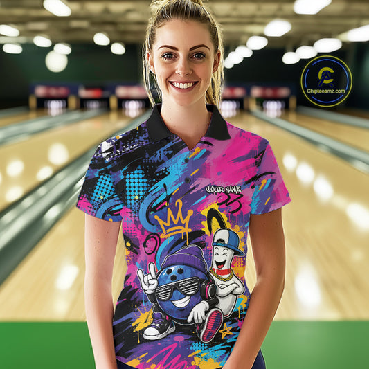 A woman stands in a bowling alley wearing a short-sleeve polo shirt with a colorful graffiti-style bowling graphic. The shirt features bright pink, blue, purple, yellow, and black splashes with cartoon bowling characters and the text 'YOUR NAME'.