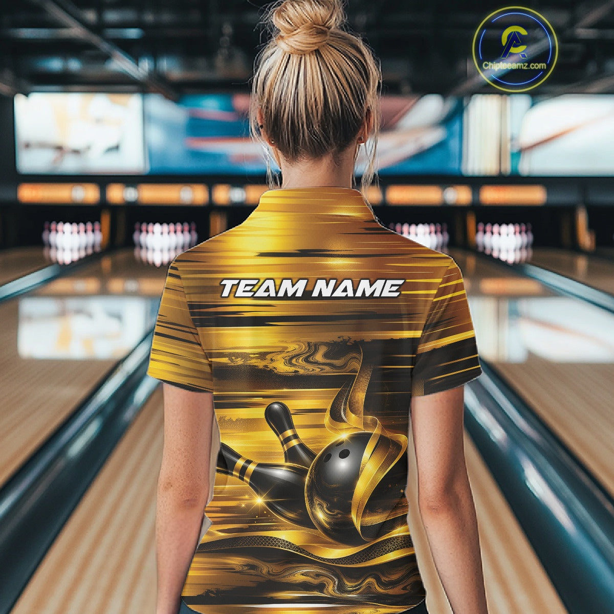 Back view of a female model at a bowling lane wearing a bowling quarter-zip shirt with glossy gold and black bowling ball and pins artwork.