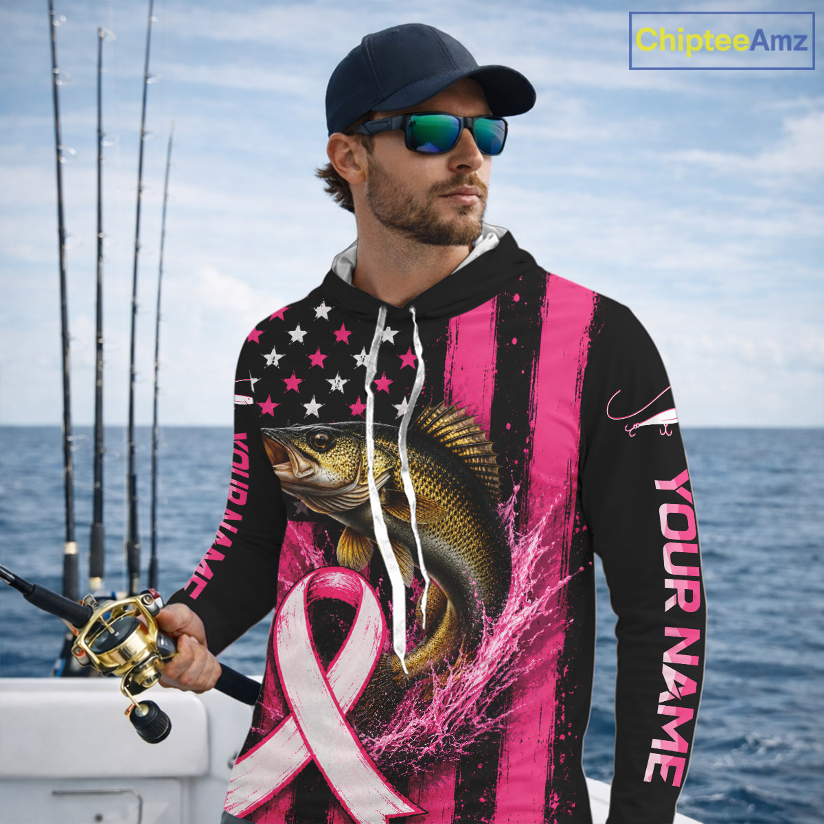 A man wearing a custom pink and black fishing hoodie with a walleye graphic, a pink ribbon, and 'YOUR NAME' printed on the sleeve, standing on a boat with fishing rods and the ocean in the background.