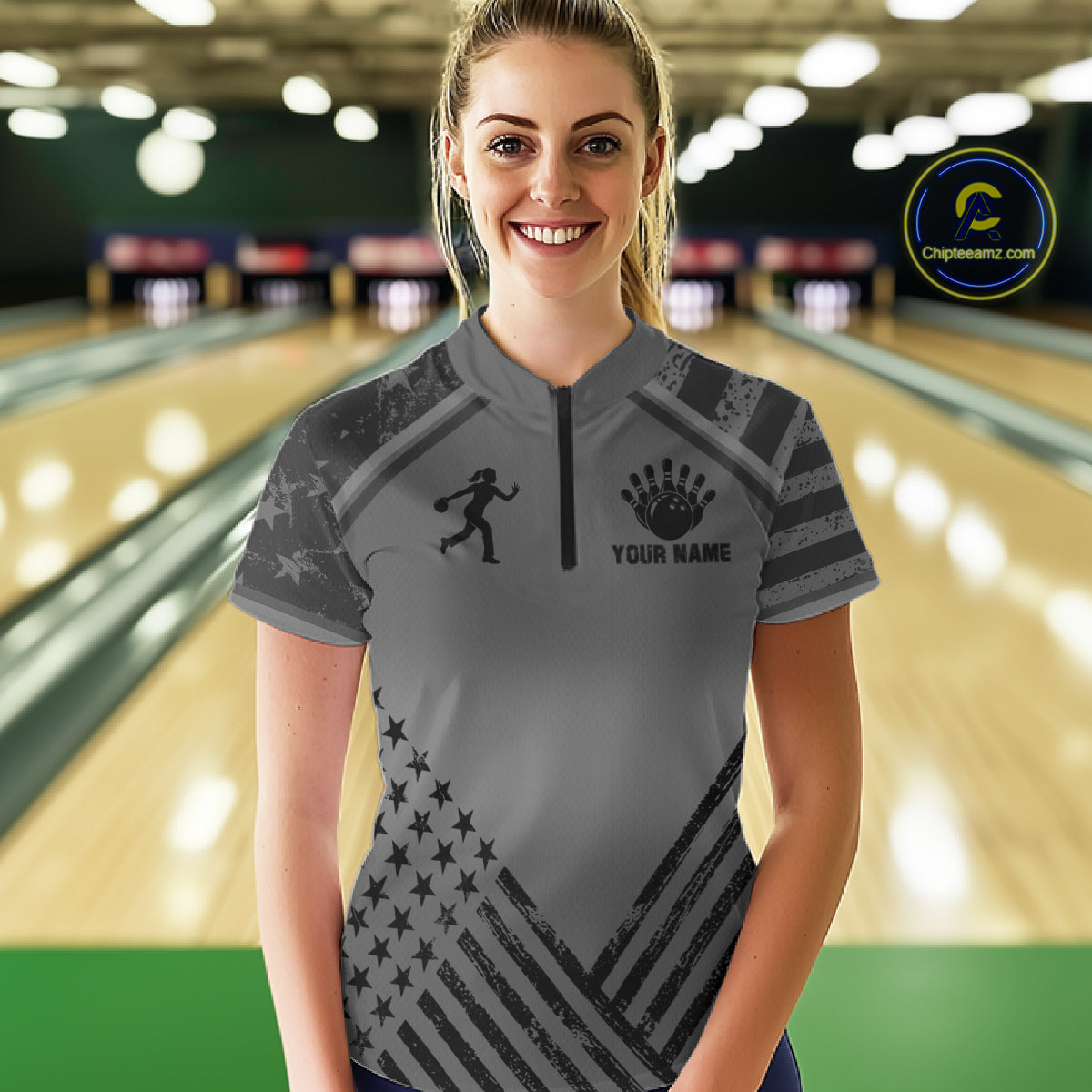 Front view of woman wearing a grey American flag bowling quarter zip shirt inside a bowling alley.